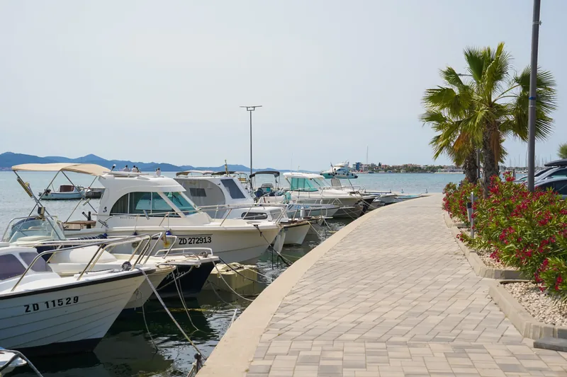 Privezane brodice uz popločanu šetnicu s palmama i oleandrima