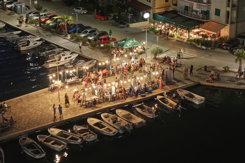 Ljetna večer na rivi: štandovi pod rasvjetom, posjetitelji i brodice u luci